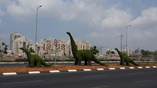 Three dinosaur-shaped topiary alongside a road with city buildings in the background